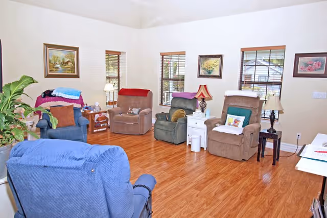 A cozy living room with five recliner chairs arranged around the room. The chairs are in various colors including blue, brown, and green, each with a blanket or pillow. The room has wooden flooring, three windows with blinds, several framed paintings on the walls, and small side tables with lamps and decorative items.