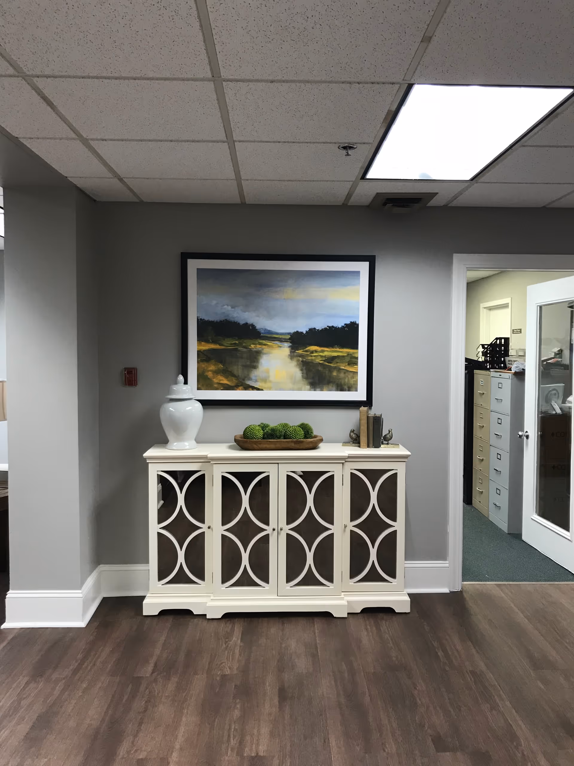 A cream decorative console table topped with a white vase and a bowl of green orbs beneath a framed landscape painting in an interior hallway.