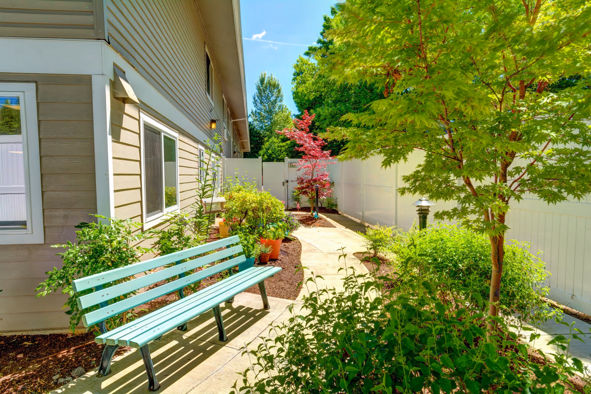 A sunny outdoor garden area at Silver Creek Senior Living featuring a light blue bench along a concrete pathway, surrounded by green shrubs, small trees, and potted plants. The area is enclosed by a white fence and adjacent to a beige building with windows.