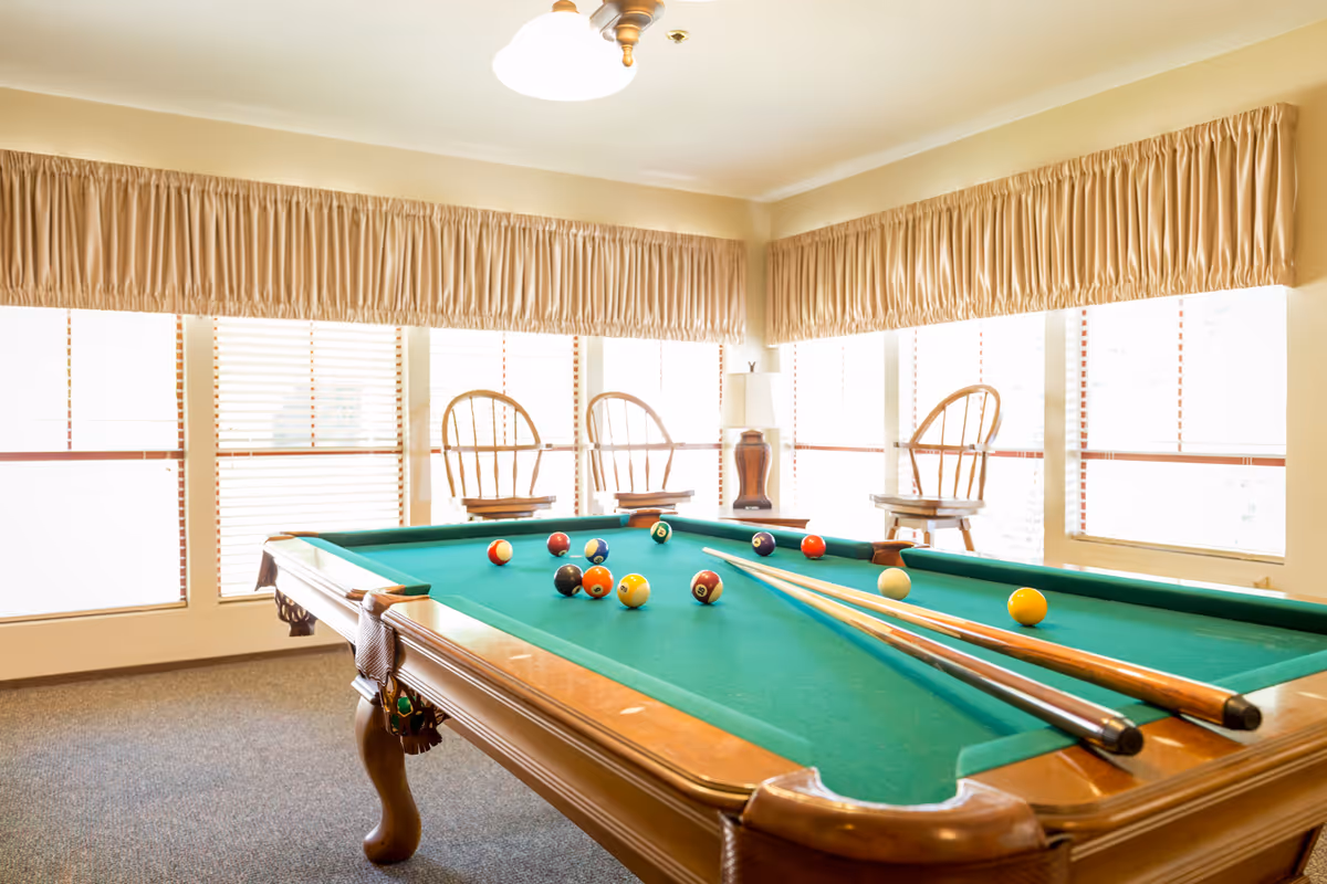 A sunlit recreation room with a green pool table, cues and balls, and chairs by large windows.