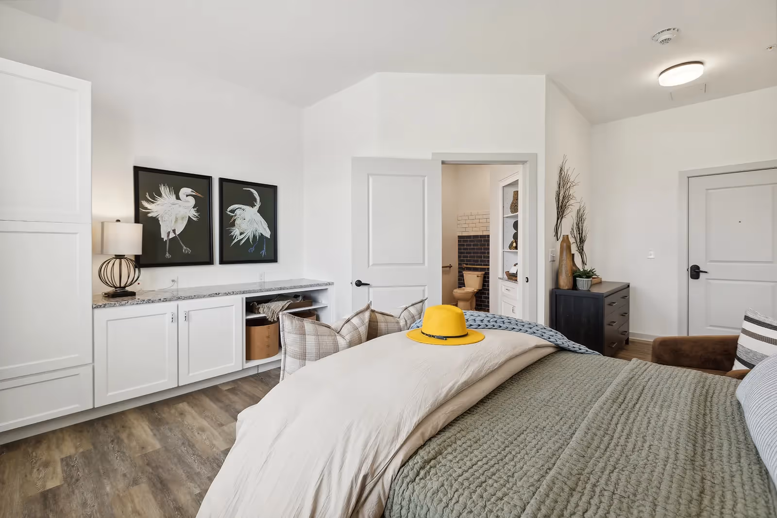 A bright bedroom with a neatly made bed featuring a green quilt and beige blanket, topped with a yellow hat. The room has white walls and wood flooring. There is a white cabinet with a granite countertop, two framed bird artworks above it, and a lamp. An open door reveals a bathroom with a toilet and tiled walls. A dark dresser with decorative plants is against the far wall.