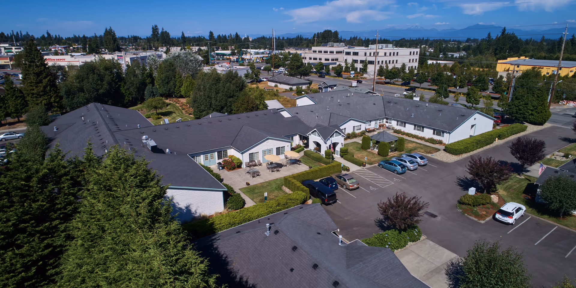 Aerial view of South Pointe Assisted Living facility showing a single-story building with a dark roof, surrounded by trees and greenery. There is a parking lot with several cars parked, and outdoor seating areas with tables and umbrellas. The background includes other buildings and a clear blue sky with some clouds.