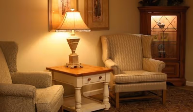 A cozy interior seating area with two upholstered armchairs flanking a wooden side table with a decorative lamp on it. Behind one chair is a wooden cabinet with glass doors displaying decorative items, and a framed artwork hangs on the wall above the table.