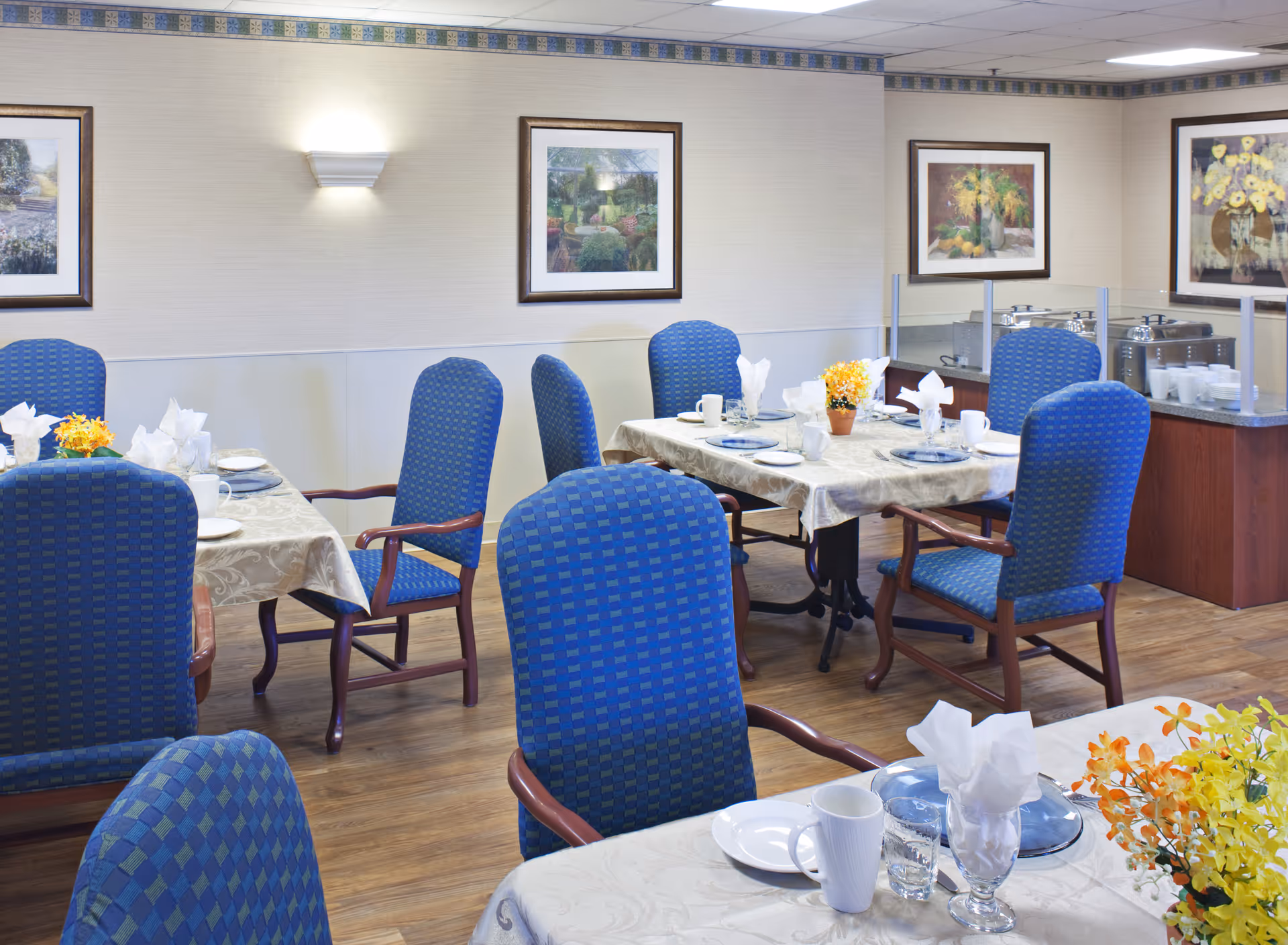 A dining room with several tables covered in white tablecloths, each set with plates, cups, glasses, and napkins. Blue upholstered chairs with wooden armrests surround the tables. The walls are decorated with framed paintings and a wall-mounted light fixture. A buffet area with covered serving dishes and cups is visible in the background.