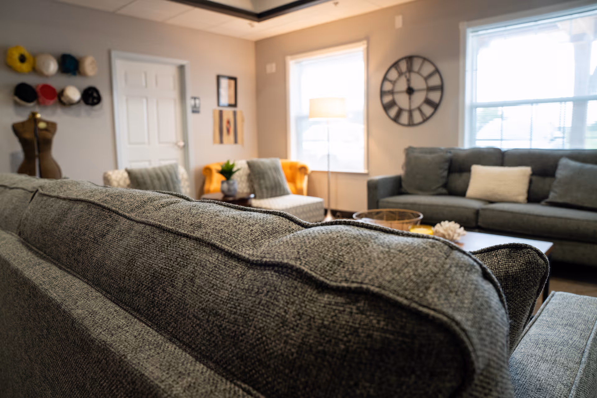 A cozy living room with gray upholstered sofas, a wooden coffee table, a yellow armchair, a floor lamp, a large wall clock, and two windows letting in natural light.