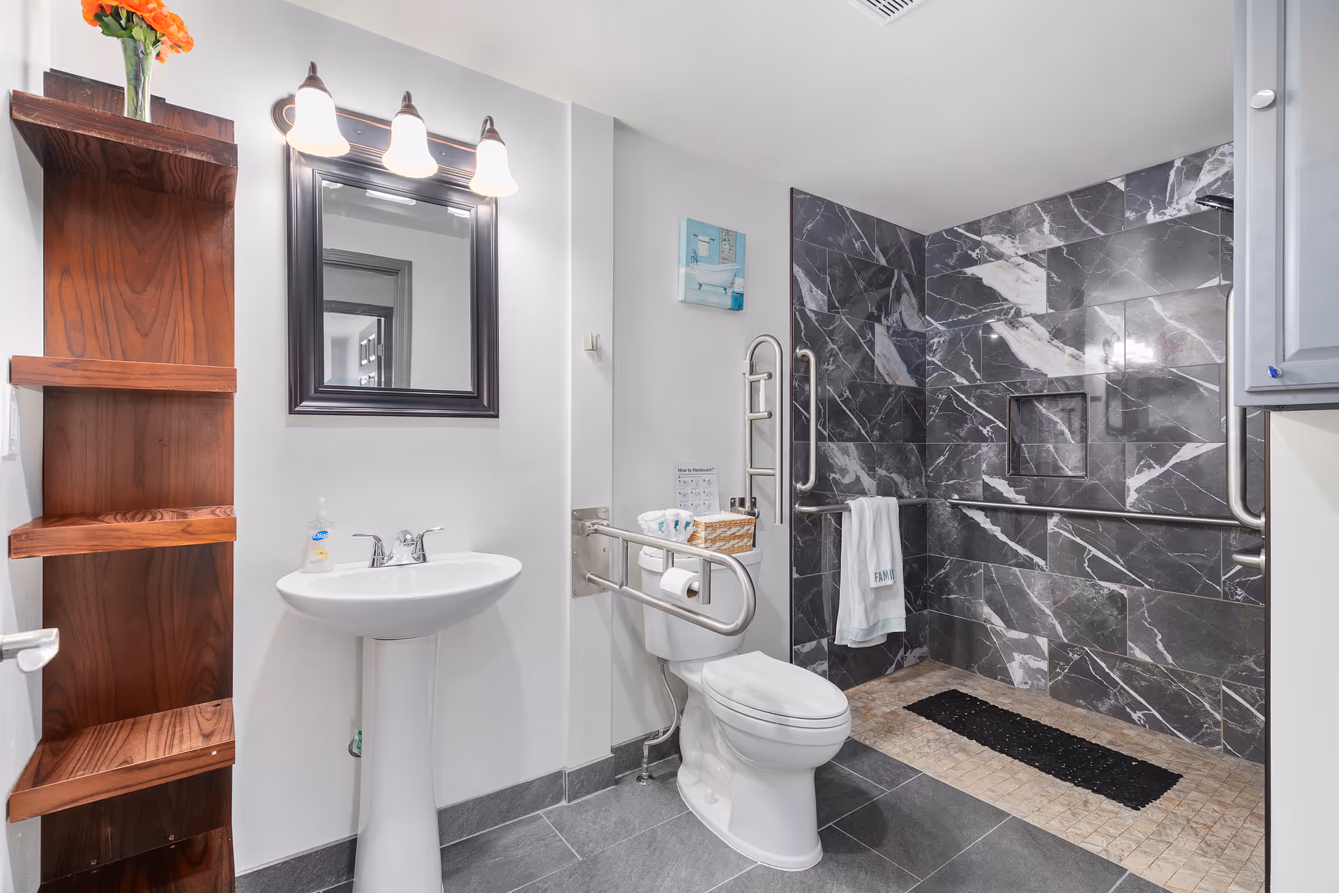 A modern bathroom with a white pedestal sink, a toilet equipped with safety grab bars, and a walk-in shower with dark gray and white marble tiles. There is a wooden shelving unit on the left wall, a mirror with three light fixtures above the sink, and a towel hanging on a grab bar near the shower.