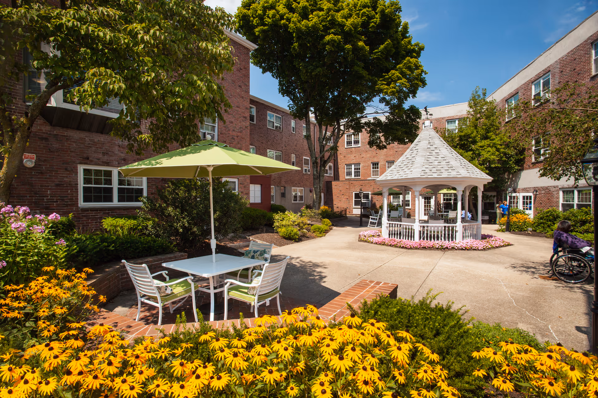 Outdoor courtyard area at Royal Health Braintree featuring a white gazebo surrounded by pink flowers, a patio table with four chairs and a green umbrella, yellow flowers in the foreground, trees, and brick buildings in the background under a clear blue sky.