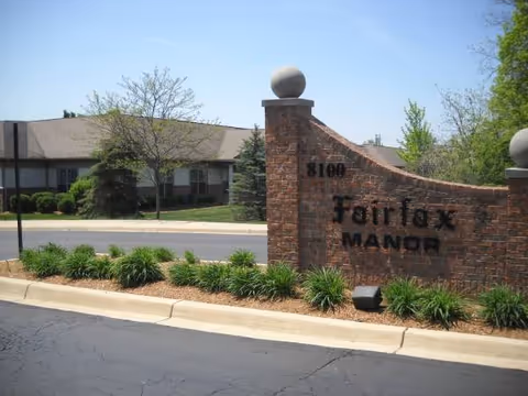 Exterior brick entrance sign for Fairfax Manor with shrubs and a driveway