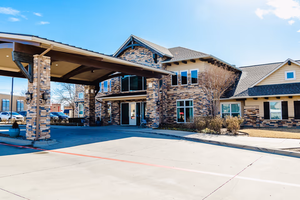 Front entrance of a stone-clad assisted living building with a covered porte-cochere and driveway under a clear blue sky.