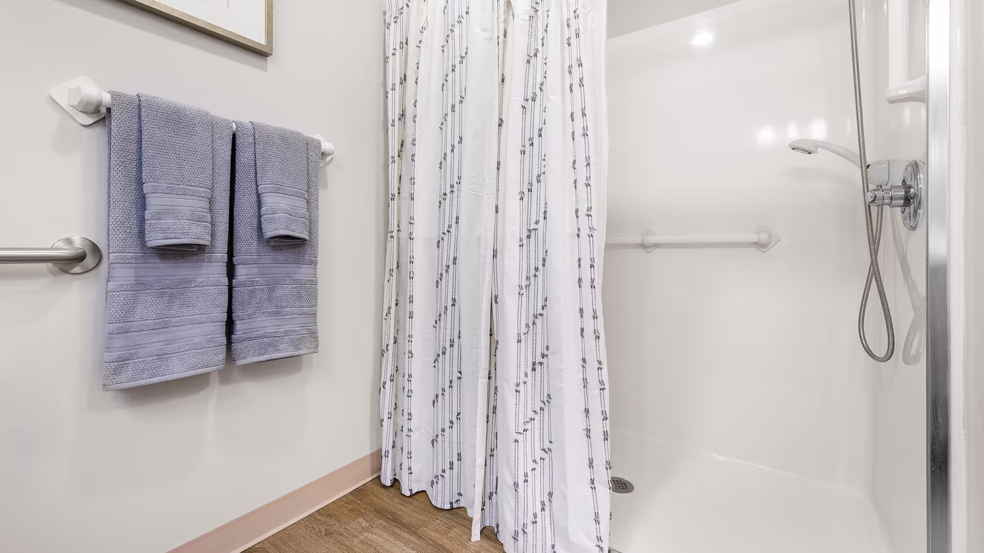 A clean bathroom shower area with a white shower curtain featuring a subtle pattern, a handheld showerhead, and a grab bar on the wall. Two folded lavender towels hang on a towel rack mounted on the wall next to the shower. The floor has a wood-like finish.