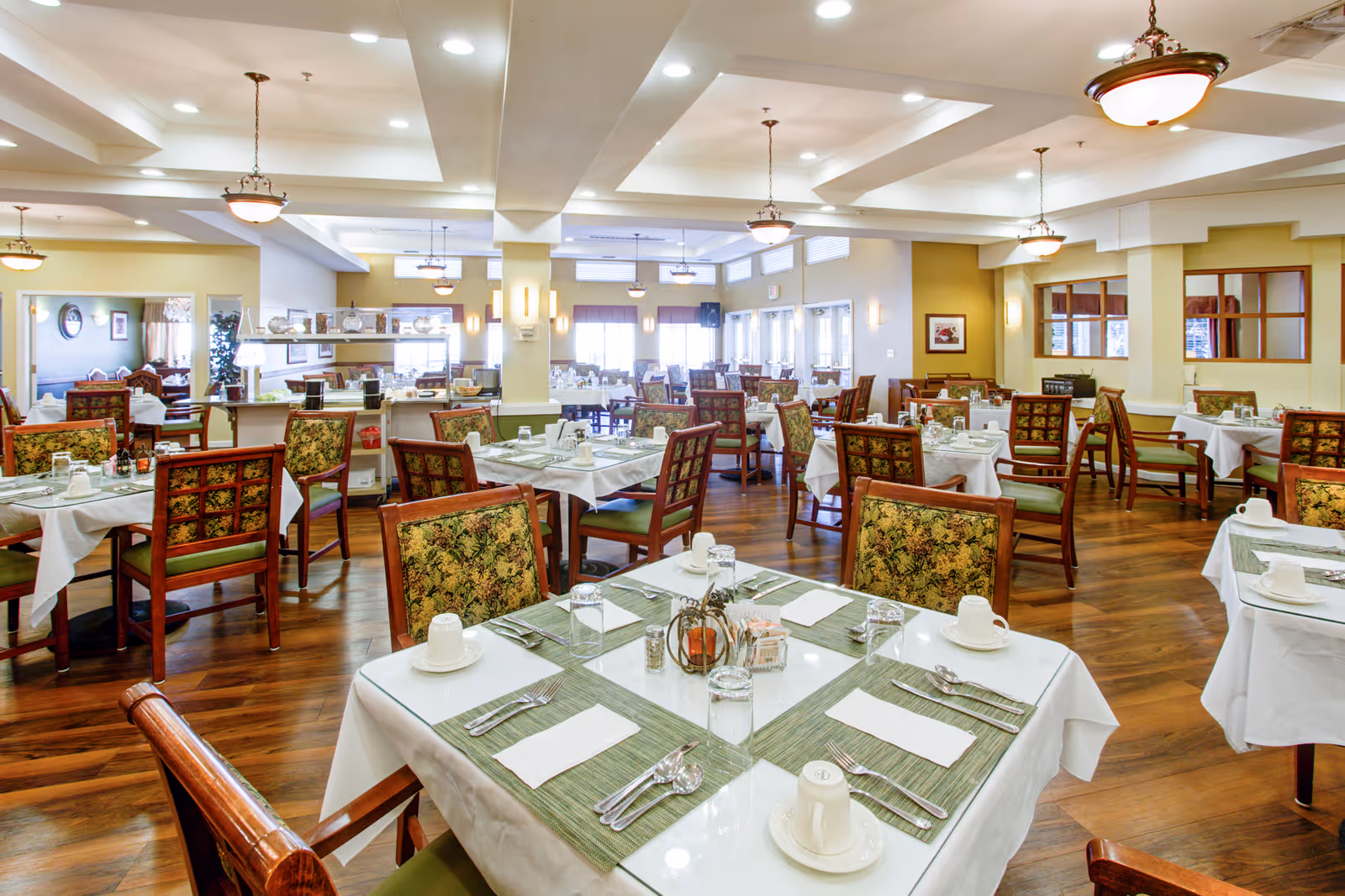 A spacious senior living dining room with multiple tables set for meals. Each table is covered with a white tablecloth and green placemats, and is set with cups, glasses, utensils, and napkins. The chairs have wooden frames with floral-patterned upholstery. The room features warm lighting with hanging light fixtures and large windows allowing natural light to enter.