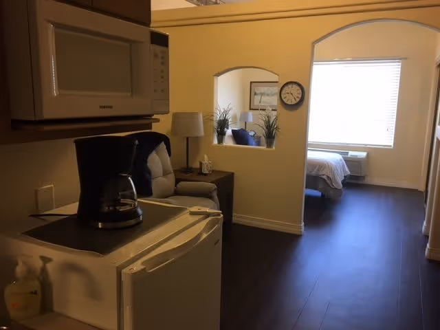 Interior view of a senior living facility room showing a small kitchenette area with a microwave, coffee maker, and mini refrigerator. Beyond the kitchenette, there is a cozy seating area with a cushioned chair and side table with a lamp. Further in the background, a bedroom area with a bed, window with blinds, and an air conditioning unit is visible.