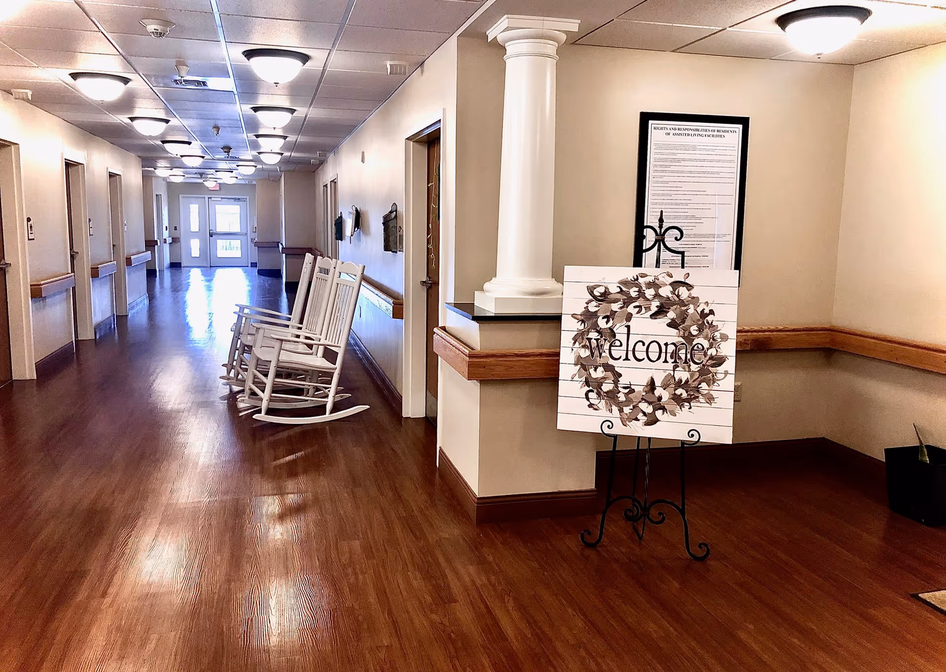 Long indoor hallway in a memory care facility with wood floors, white rocking chairs, and a decorative 'welcome' sign on an easel by a column.