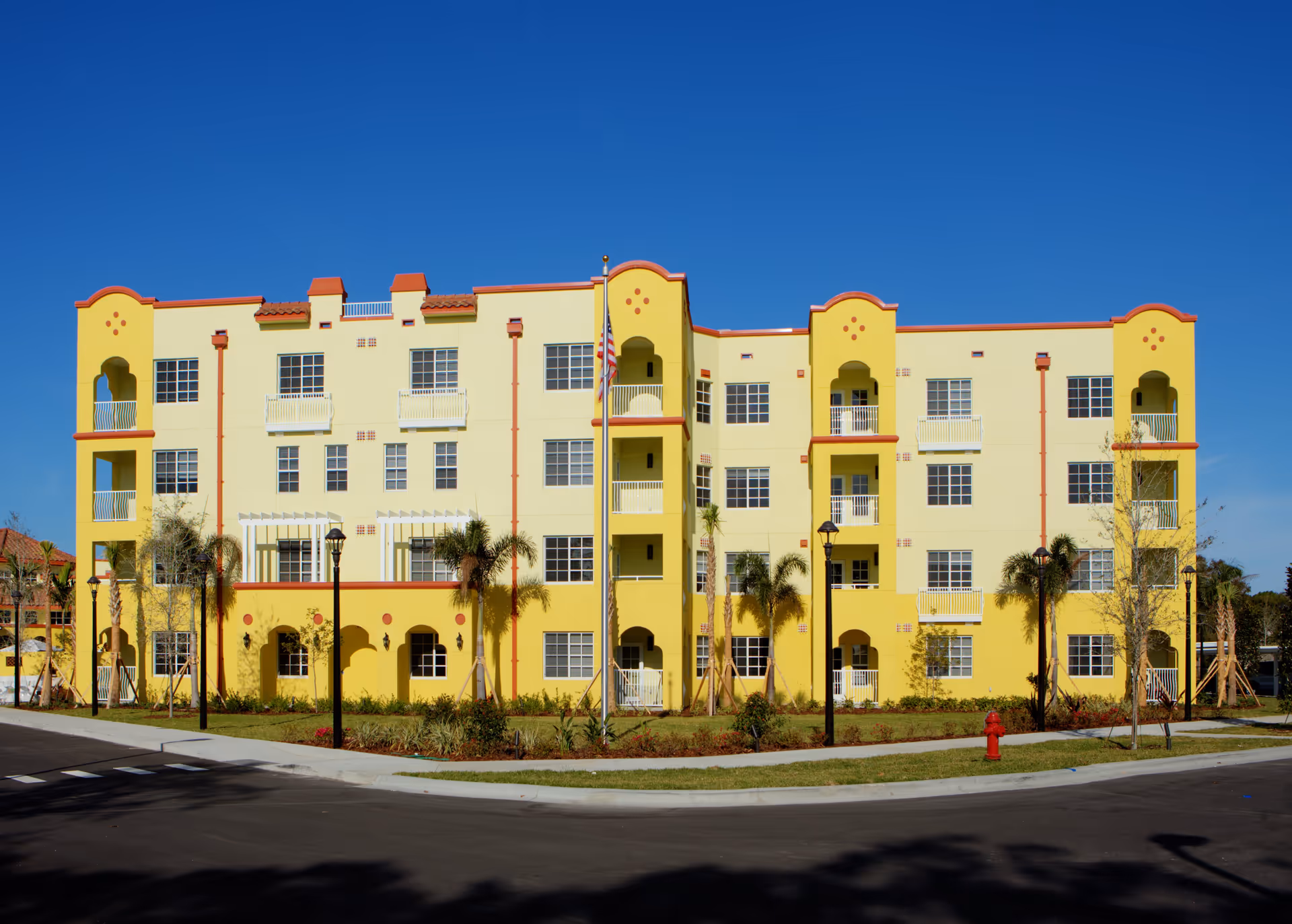 Front exterior view of a four-story yellow and orange building with multiple windows and balconies, surrounded by palm trees and street lamps under a clear blue sky.