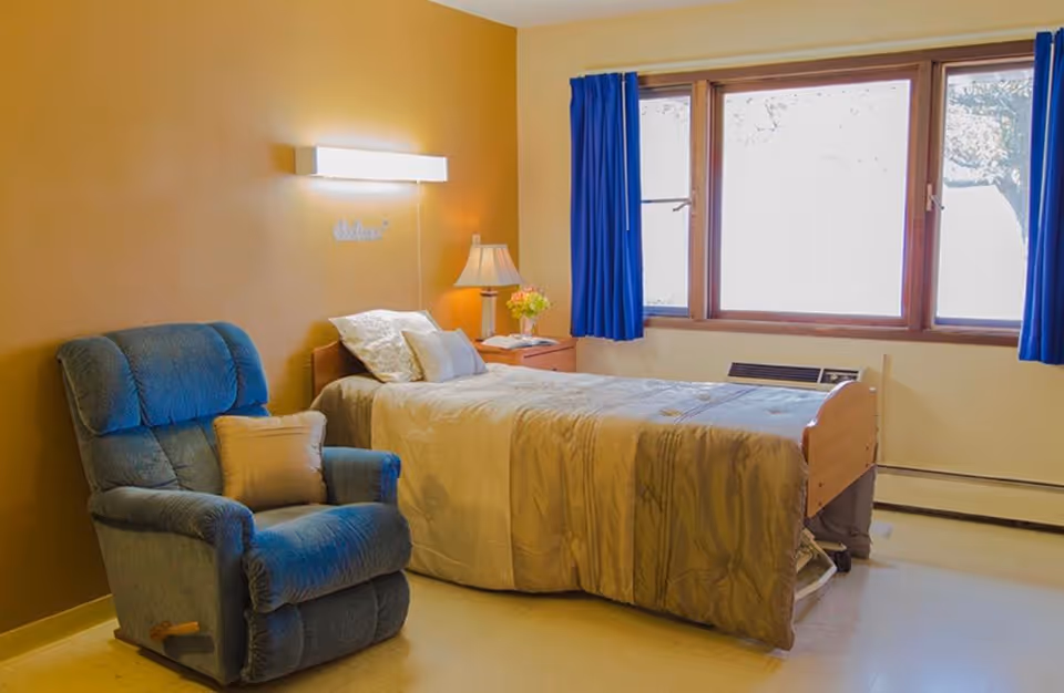 A cozy nursing facility bedroom with a single bed covered in a beige comforter, a blue recliner chair with a pillow, a wooden nightstand with a lamp and flowers, and a large window with blue curtains letting in natural light.