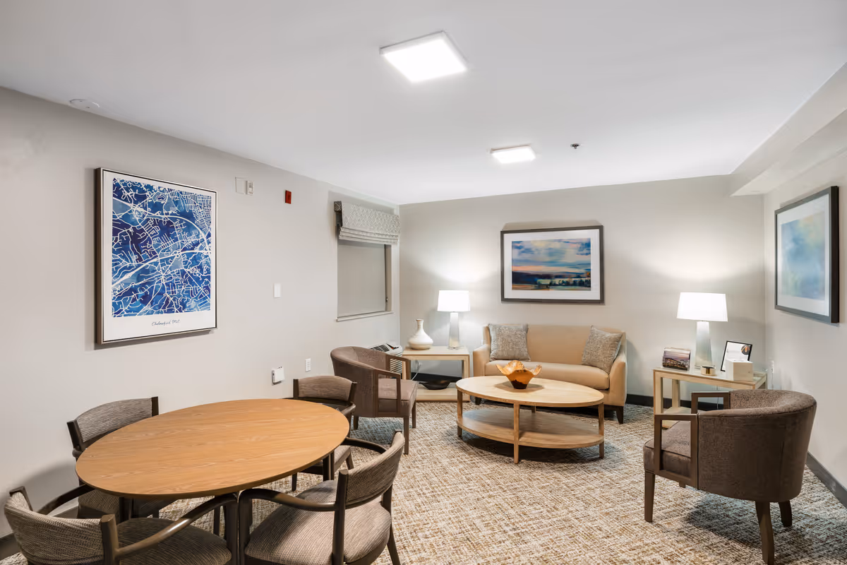 A cozy senior living community lounge featuring a beige sofa with two cushions, two brown armchairs, a round wooden coffee table, and a round wooden dining table with six chairs. The room is softly lit by two table lamps on side tables and overhead ceiling lights. The walls are decorated with framed artwork, including a blue map and landscape paintings. The floor is carpeted in a neutral pattern.