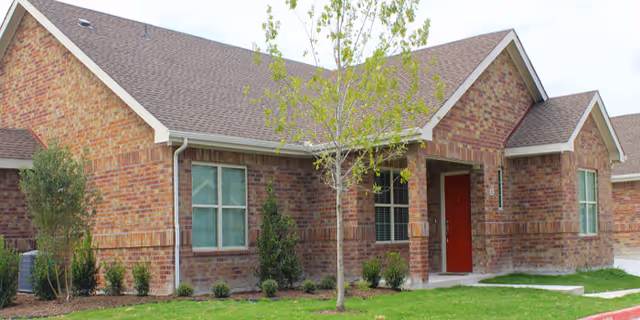 Exterior view of a single-story brick building with a pitched roof, several windows, a red front door, and a small tree and shrubs in the front yard.