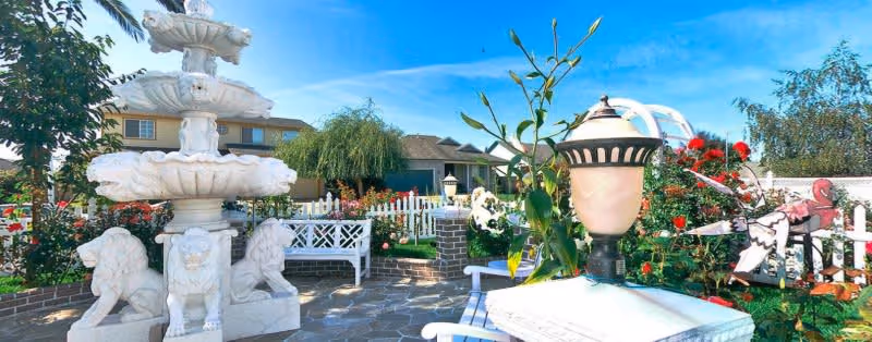 Outdoor garden area at a senior care facility featuring a large white multi-tiered fountain with lion sculptures at its base, white benches, a white picket fence, various flowering plants including red roses, and a decorative lamp post under a clear blue sky.