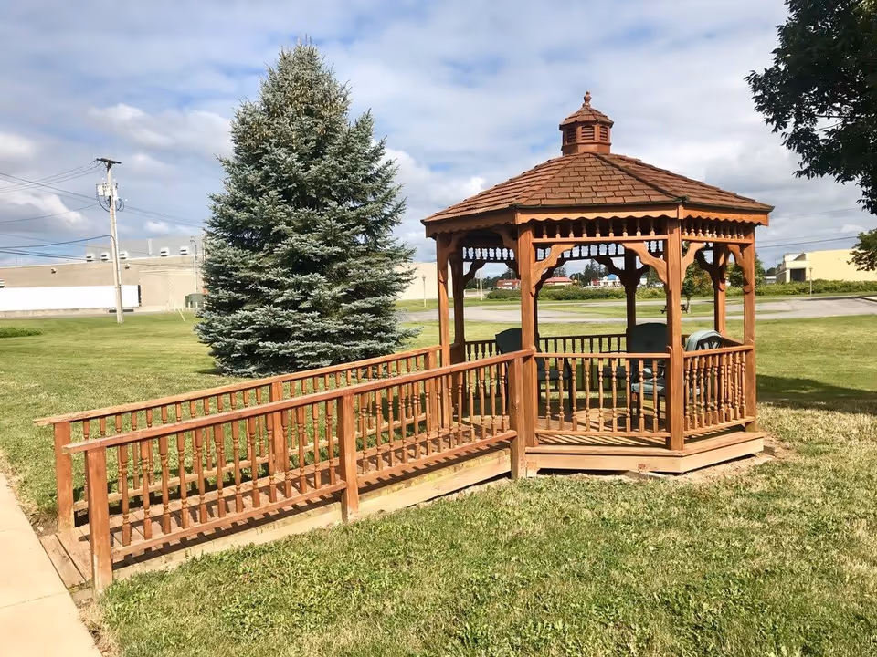 A wooden gazebo with a shingled roof and decorative railings, situated on a grassy area with a large evergreen tree nearby. The gazebo has a wooden ramp leading up to it and contains several chairs inside. The sky is partly cloudy and there are buildings and power lines in the background.