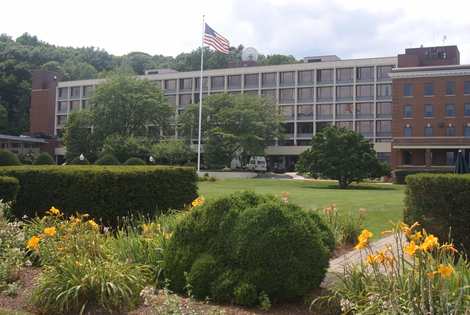 Exterior view of Masonicare Health Center showing a multi-story building with large windows, surrounded by green trees, bushes, and a well-maintained garden with yellow flowers. An American flag flies on a flagpole in front of the building under a partly cloudy sky.