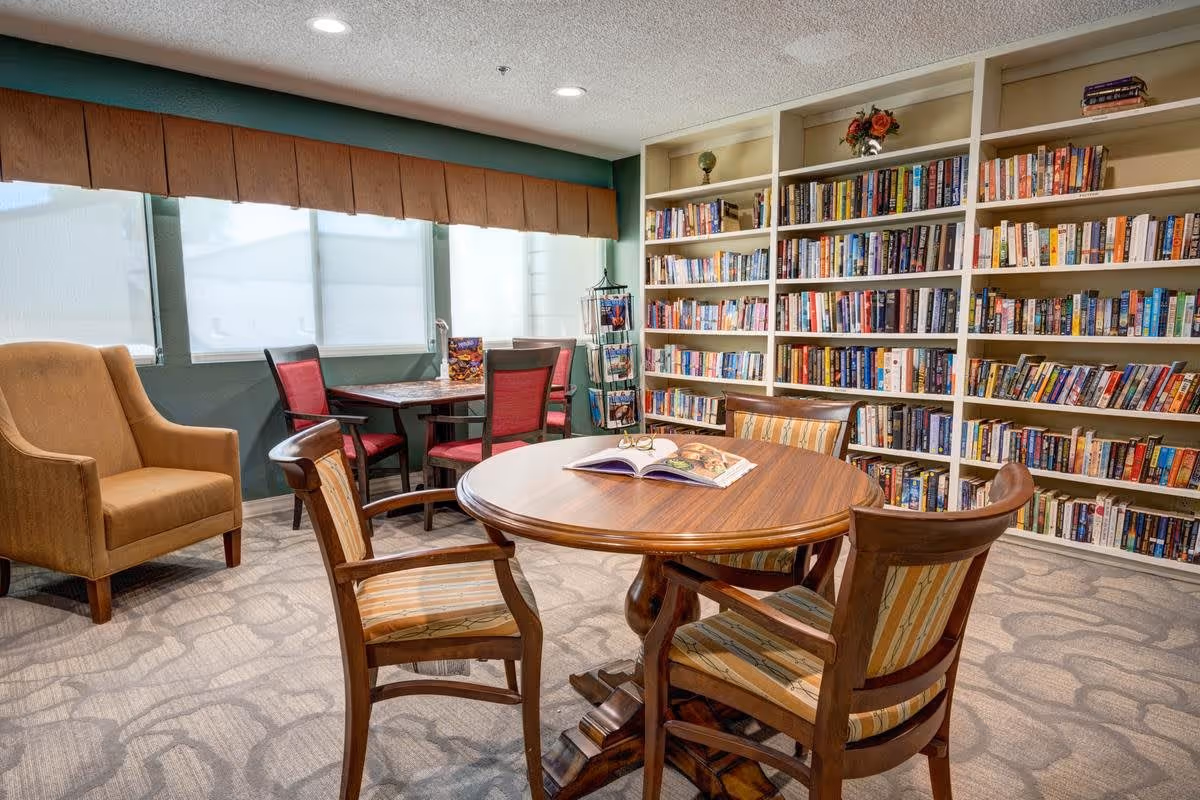 A cozy reading room with a large bookshelf filled with books along one wall. There is a round wooden table with four wooden chairs featuring striped cushions in the center of the room. A book and a pair of glasses are placed on the table. In the background, there is a smaller table with four red cushioned chairs near windows with blinds, and a comfortable beige armchair is positioned to the left.