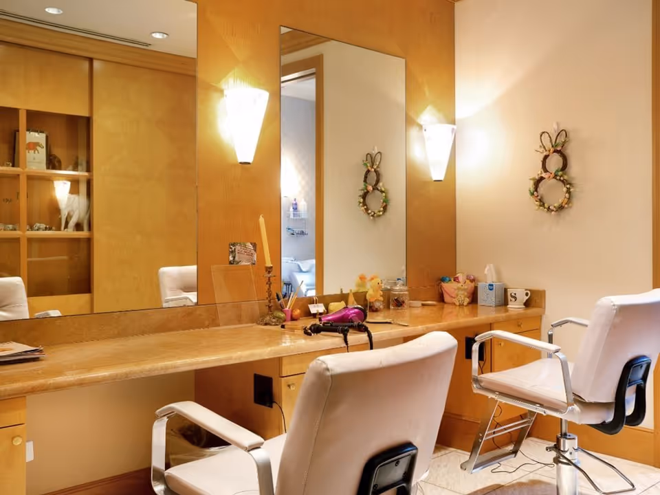 Well-lit salon vanity area with two white styling chairs, a large mirror and a countertop holding a hair dryer and decorative items.