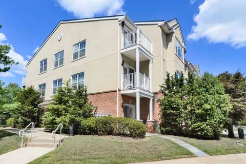 Three-story beige Horizons at Franklin Lakes senior living building with balconies, brick base, shrubs, and sidewalk.