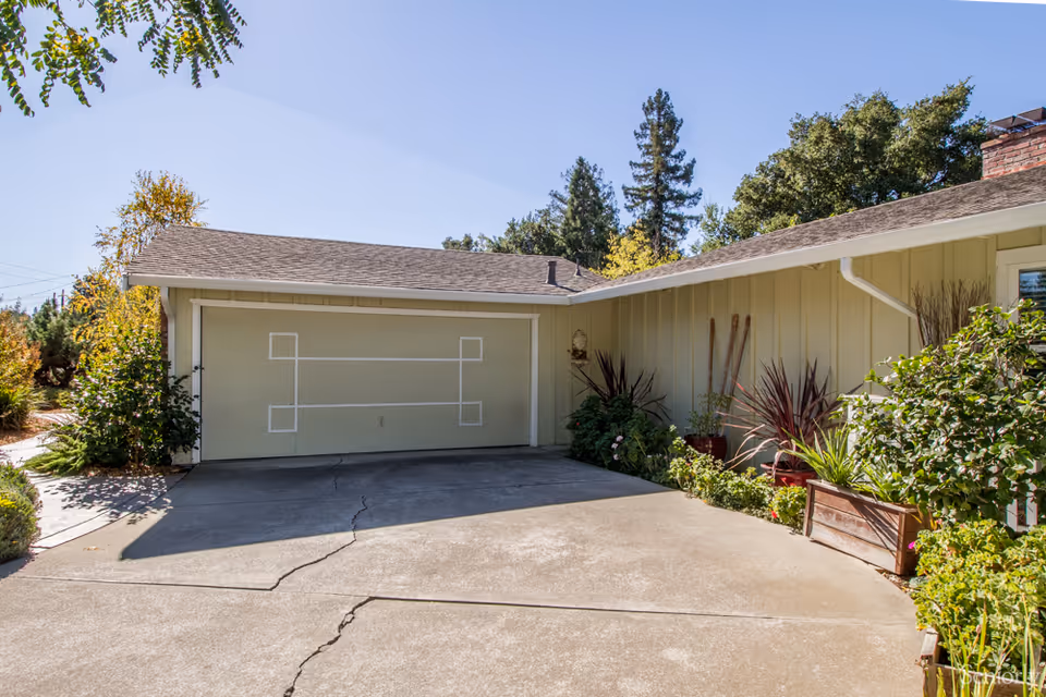 Driveway leading to a closed garage door of a single-story house with beige siding, surrounded by green plants and trees under a clear blue sky.