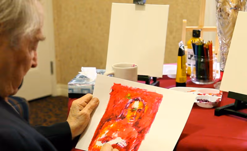 An elderly person holding and looking at a colorful painting of a person, seated at a table with art supplies including paint tubes, colored pencils, and blank canvases on easels in the background.