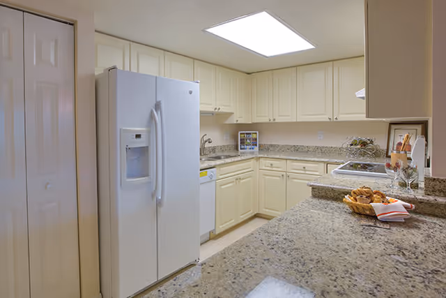 Well-lit kitchen with cream cabinets, granite countertops, a white side-by-side refrigerator, sink, and a breakfast bar with pastries.