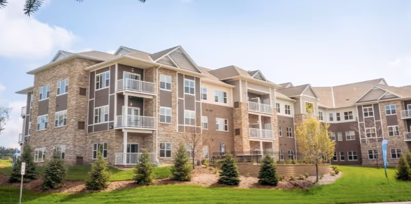 Exterior view of a multi-story senior living facility building with balconies, large windows, and a landscaped lawn with small trees and shrubs under a partly cloudy sky.