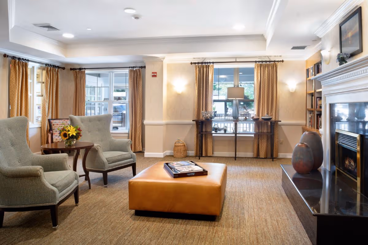 A cozy living room area with two gray upholstered armchairs, a round wooden side table with a vase of sunflowers, a large tan leather ottoman with a tray on top, and a fireplace with decorative vases on the mantel. The room has beige walls, carpeted floor, large windows with gold curtains, a console table with a lamp and decorative items, and built-in bookshelves.