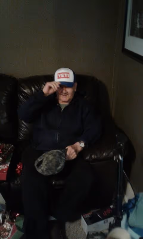 An elderly man sitting on a dark brown leather couch in a dimly lit room. He is wearing a dark jacket and a white and blue YETI cap, which he is adjusting with one hand. There is a camouflage hat on his lap and various items, including a box and some wrapped gifts, are on the floor nearby. A framed picture is partially visible on the wall to the right.