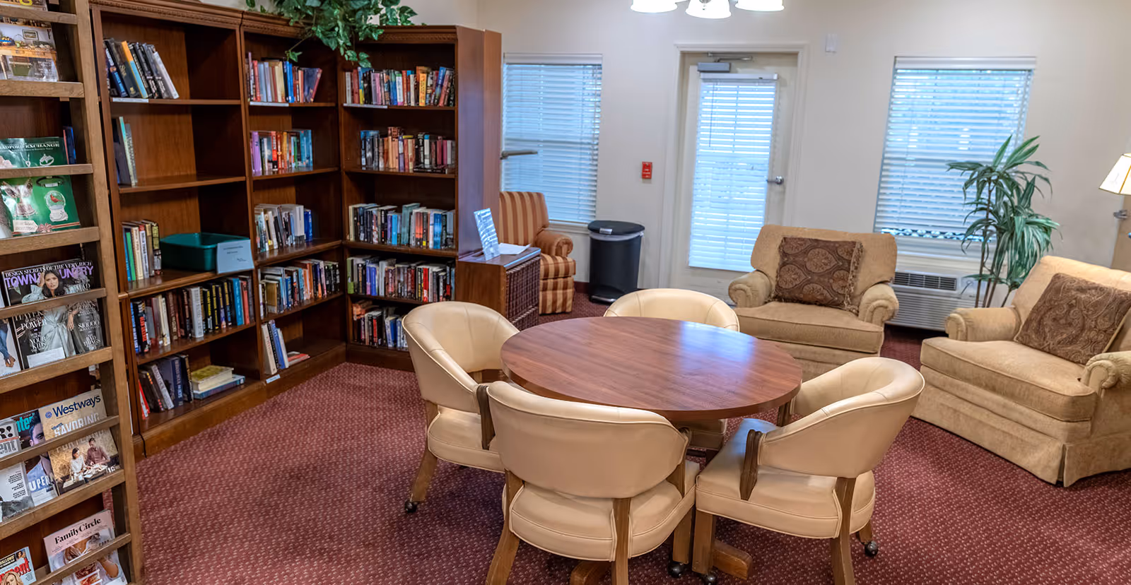 Cozy communal lounge with a round wooden table surrounded by padded chairs, bookshelves and armchairs.
