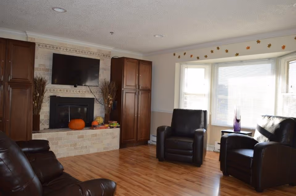 Bright living room with hardwood floors, leather armchairs, a wall-mounted TV above a fireplace flanked by cabinets, and a bay window.