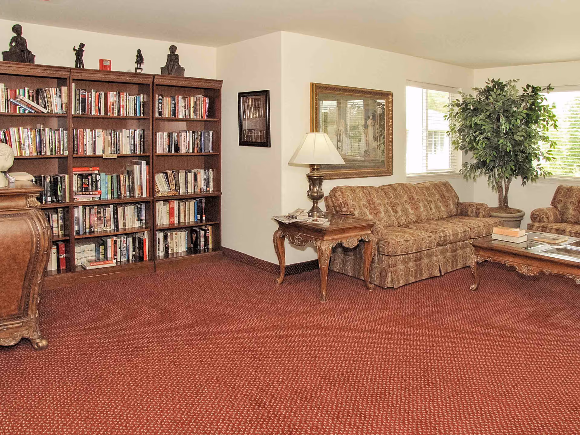 Cozy sitting room with bookshelves along the left wall, a patterned sofa, side and coffee tables, a lamp, and a potted plant by the windows.