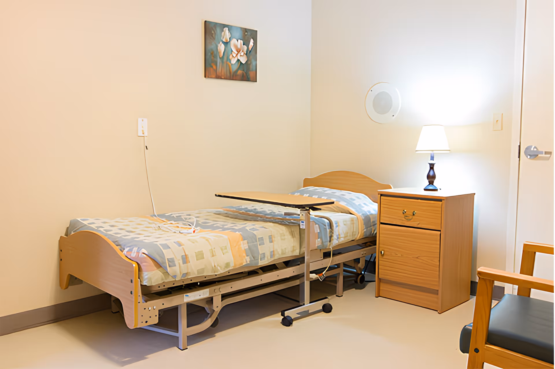 A simple senior care bedroom with a single hospital-style bed covered with a patterned blanket, a small adjustable overbed table, a wooden nightstand with a lamp, a wooden chair with a dark cushion, and a floral painting on the wall.