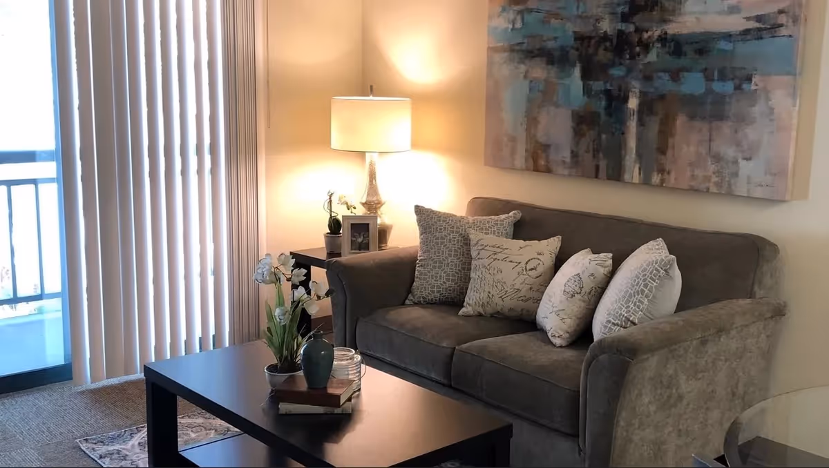 A cozy living room with a gray sofa adorned with five decorative pillows. A black coffee table in front of the sofa holds a small plant, a vase, and some books. A side table next to the sofa has a lamp, a small plant, and a framed photo. Vertical blinds cover a sliding glass door leading to a balcony. A large abstract painting hangs on the wall above the sofa.