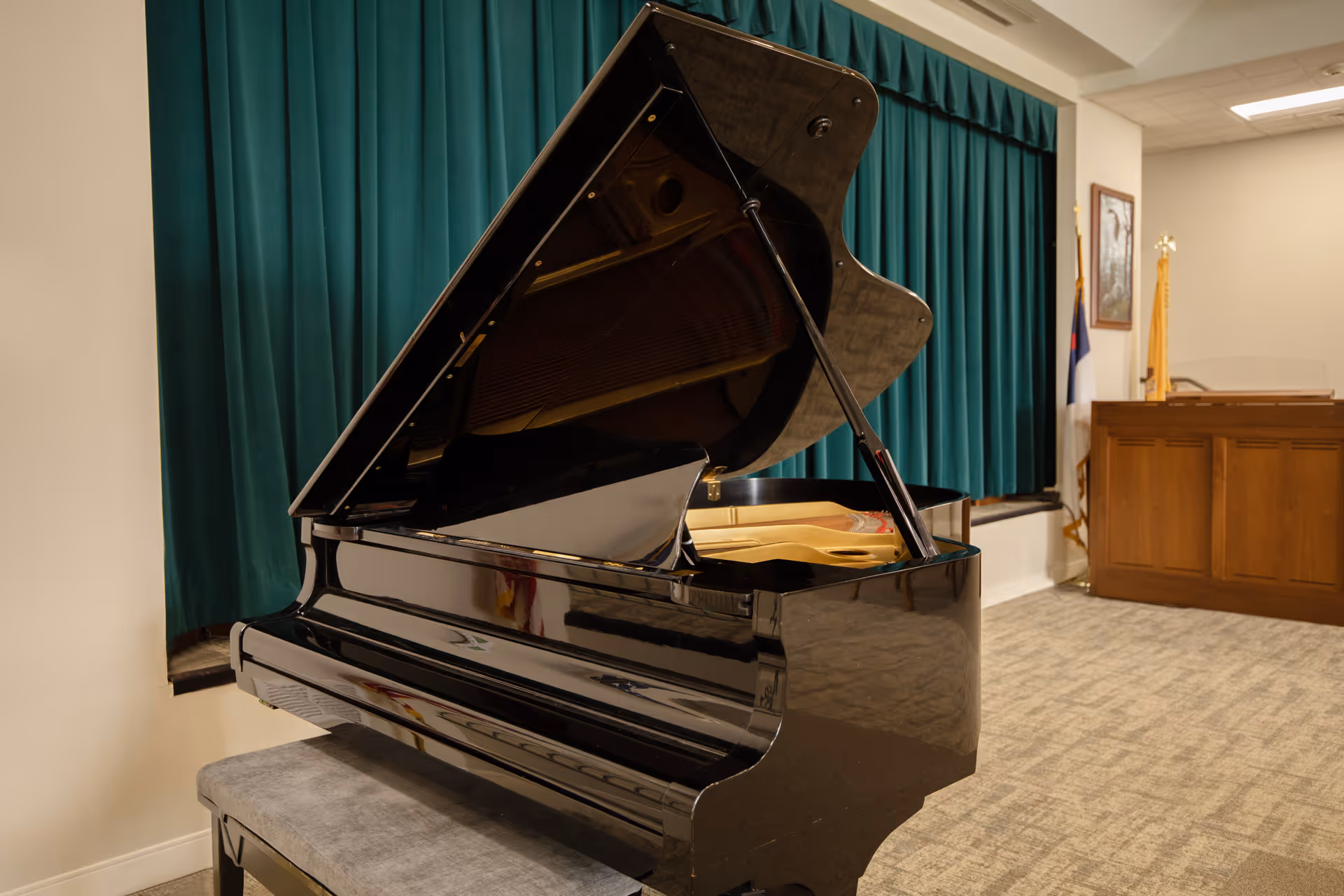A black grand piano with its lid open in a room with beige carpet and walls. Behind the piano is a green curtain covering a window, and to the right is a wooden podium with flags and framed pictures on the wall.