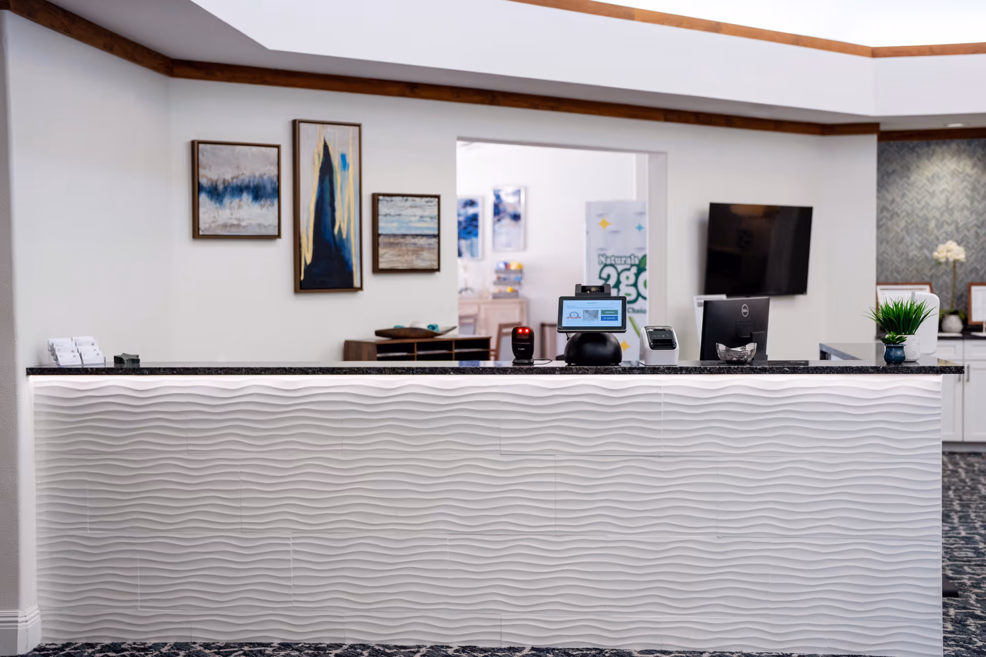 Reception desk area with a textured white front panel and a black countertop. On the countertop are a computer monitor, a payment terminal, a barcode scanner, and a small potted plant. Behind the desk is a wall with three framed abstract paintings and an open doorway leading to another room with more artwork and furniture.