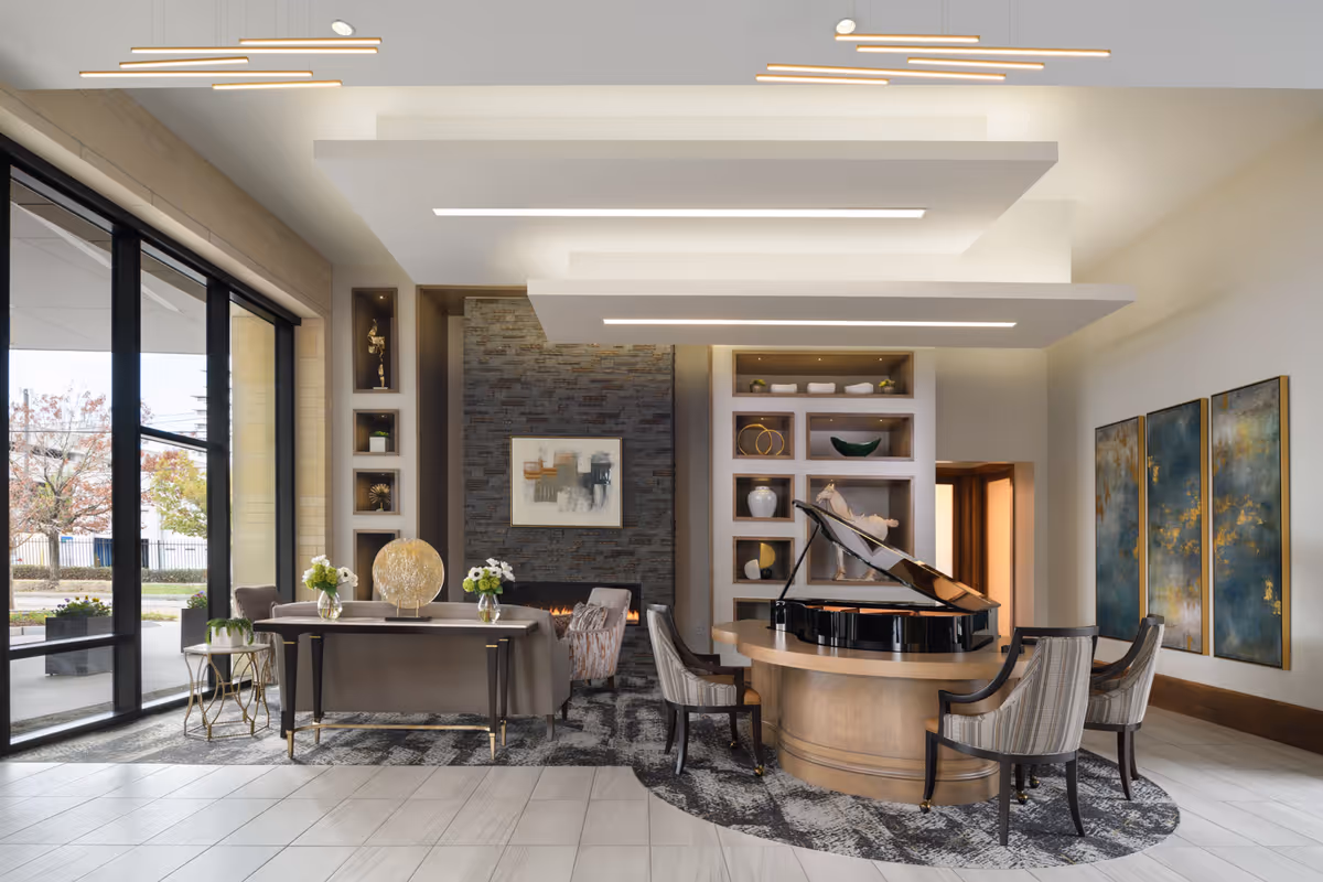 A modern senior living facility common area featuring a grand piano on a round wooden table surrounded by four upholstered chairs. The room has large floor-to-ceiling windows on the left side letting in natural light, a stone accent wall with a framed abstract painting, built-in shelves with decorative items, and three large abstract paintings on the right wall. The ceiling has layered rectangular light fixtures, and there are small tables with flower arrangements near a sofa and armchairs.