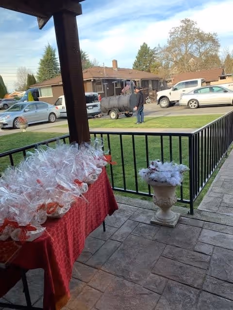 A covered patio area with a red tablecloth-covered table holding multiple wrapped gift baskets. A white decorative planter with white flowers is placed near the black metal railing. In the background, there is a grassy lawn, several parked cars, a person standing near a large barbecue smoker, and residential buildings with trees under a blue sky.