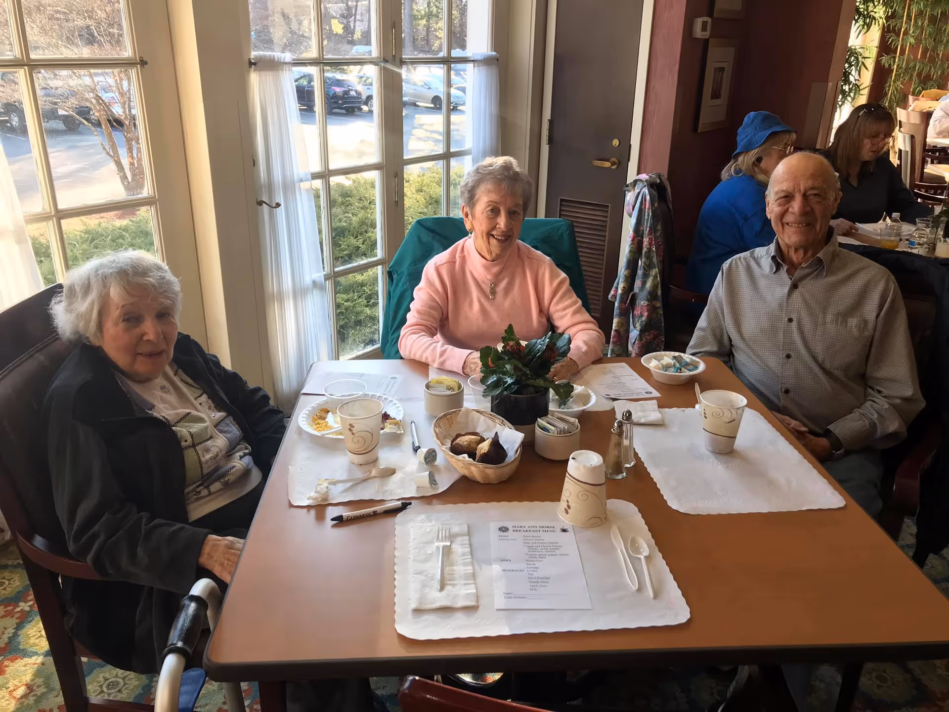 Three elderly people sitting around a dining table in a well-lit room with large windows. The table has paper placemats, disposable cups, a small plant centerpiece, and some food items. Two women and one man are smiling at the camera.