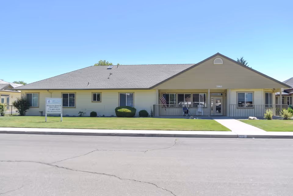 Single-story beige building with a gray roof, a small front porch with rocking chairs, an American flag, and a well-maintained lawn. A sign in front reads 'Skyline Estates Residence House & Main Office' with a phone number and address. The building number 2861 is visible above the entrance.