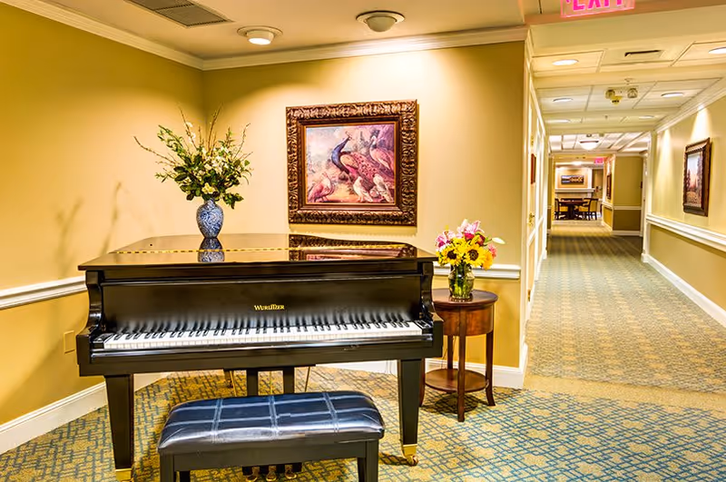 A black Wurlitzer piano with a matching bench sits in a well-lit hallway with yellow walls and patterned carpet. On top of the piano is a blue vase with flowers, and next to it is a small round wooden table with a bouquet of flowers. A framed painting of peacocks hangs on the wall above the piano. The hallway extends into the distance with more framed artwork and an exit sign visible.