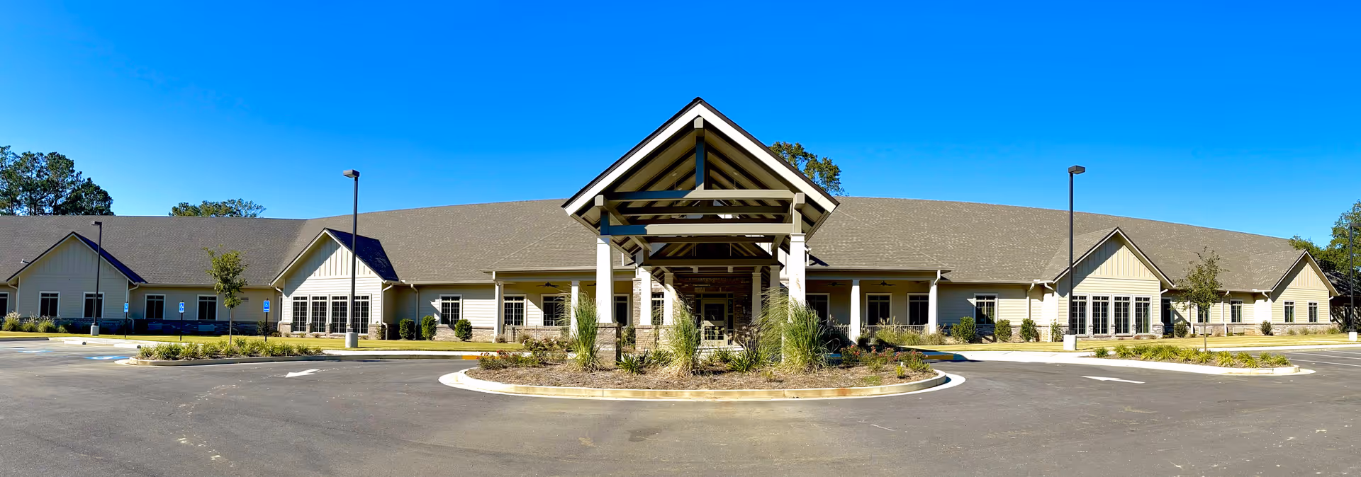 Wide exterior view of a single-story senior living facility building with a large covered entrance, surrounded by a circular driveway and landscaped greenery under a clear blue sky.