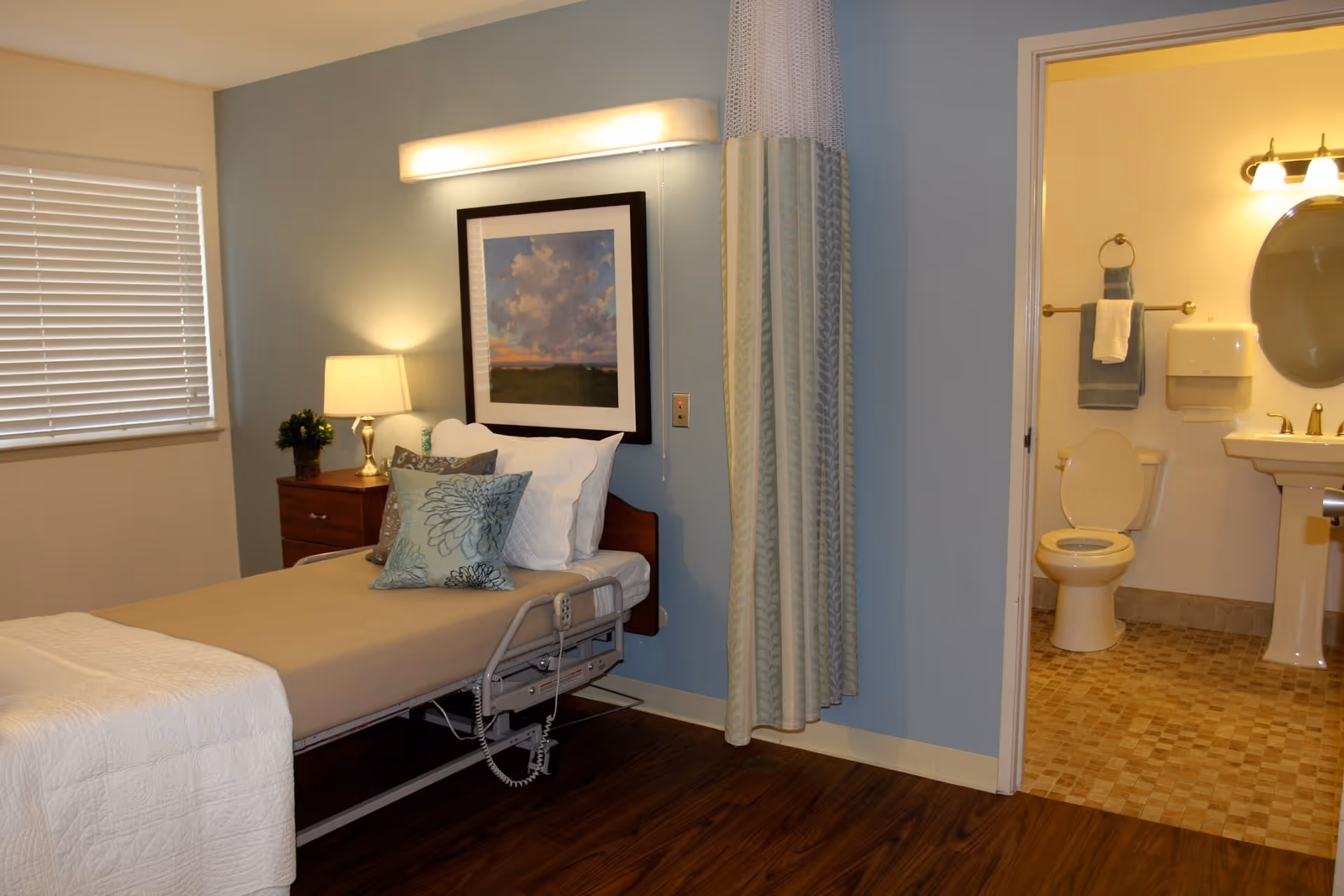 A hospital-style bed with beige bedding and decorative pillows in a room with light blue walls. Next to the bed is a wooden nightstand with a lamp and a small plant. A framed picture hangs above the bed. To the right, there is an open doorway leading to a bathroom with a toilet, sink, mirror, and towel racks.