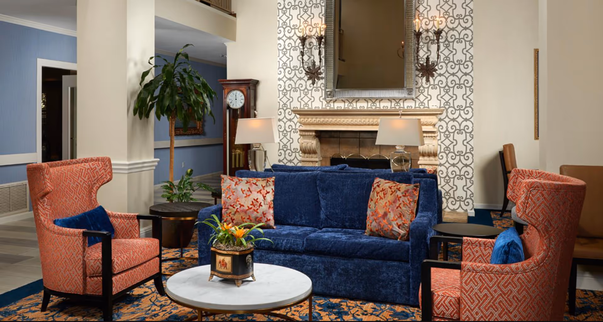 Cozy lounge area with a blue velvet sofa, two patterned armchairs, a round coffee table and a decorative fireplace with mirror.