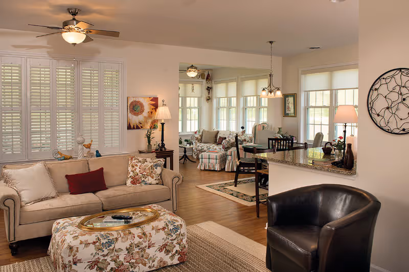 A cozy living room area with a beige sofa adorned with cushions, a floral patterned ottoman, a black leather chair, and a ceiling fan with a light. The room opens into a dining area with a table and chairs, large windows with blinds, and soft lighting from lamps and a hanging light fixture. The walls are decorated with artwork and a circular metal wall decor.