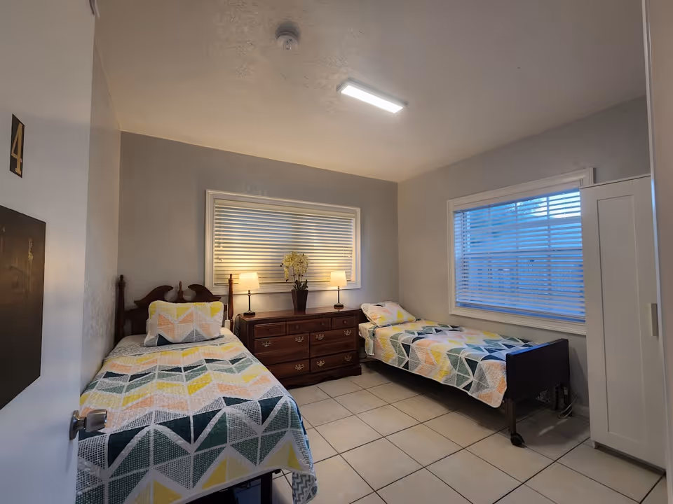 A small bedroom with two single beds, each covered with a colorful geometric patterned quilt. Between the beds is a wooden dresser with two lit table lamps and a potted plant with flowers. The room has tiled flooring, two windows with white blinds, and a white wardrobe on the right side. The door to the room is partially open, showing the number 4 on it.